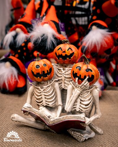 Trois squelettes avec têtes de citrouilles lisent ensemble un livre sur fond de décorations festives.