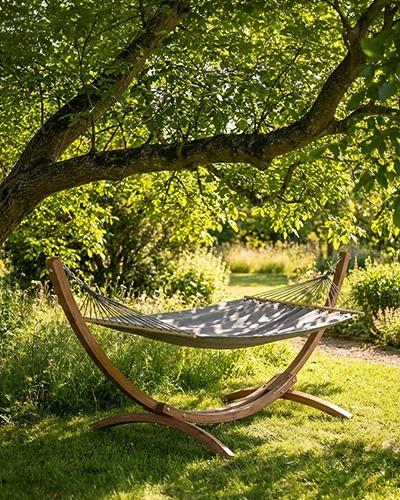 Un hamac gris sur un support en bois courbé installé sous un grand arbre dans un jardin ensoleillé.