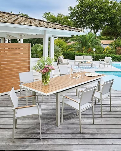 Table de jardin rectangulaire en bois et métal avec chaises assorties sur une terrasse pavée devant une maison en pierre.